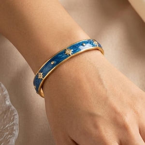 Puede incluir: Un brazalete dorado con una banda de esmalte azul. El esmalte azul está decorado con formas de estrella doradas. El brazalete se lleva en la muñeca.