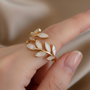 Op de afbeelding: Een goudkleurige ring met een bladerenontwerp. De ring heeft witte, ovaalvormige accenten die op bladeren lijken. De ring wordt aan een vinger gedragen en toont zijn delicate en elegante ontwerp. De ring is een sieraad.