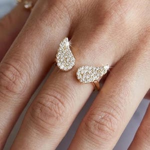 Könnte beinhalten: Ein goldfarbener Ring mit zwei tropfenförmigen Abschnitten, die jeweils mit kleinen, funkelnden, klaren Steinen besetzt sind. Der Ring wird an einem Finger getragen und zeigt das aufwendige Design und den Glanz der Steine.