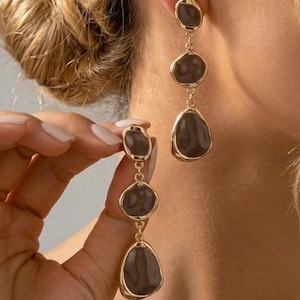 May include: A pair of long drop earrings featuring three graduated, irregular, pebble-shaped dark gray resin stones encased in polished gold-tone metal frames. The stones are linked vertically by small gold-tone metal rings, creating a dangling design.