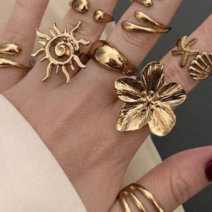 Könnte beinhalten: Eine Sammlung goldfarbener Ringe in verschiedenen Designs, darunter ein Sonnenaufgang, eine Blume, eine Muschel und ein Seestern. Die Ringe werden auf einer Hand mit burgunderfarbenem Nagellack präsentiert.