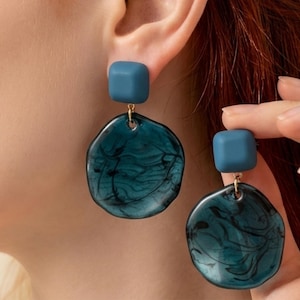 May include: A pair of teal and blue earrings. Each earring features a small, square stud in teal, connected to a larger, irregular-shaped pendant with a swirling, marbled teal design. The earrings are shown on an ear.