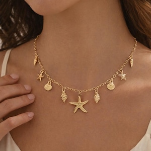 Puede incluir: Collar dorado con colgantes de conchas marinas y estrellas de mar. El collar tiene una cadena delicada y múltiples colgantes, incluyendo estrellas de mar, conchas y otros colgantes con temática marina. El collar se lleva alrededor del cuello.
