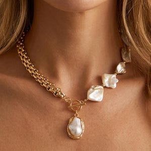 Könnte beinhalten: Eine goldfarbene Kettenhalskette mit einer Reihe unregelmäßiger, glänzender weißer Perlen auf der einen Seite und einer dicken, ineinandergreifenden goldfarbenen Kette auf der anderen. Die Kette hat einen zentralen Knebelverschluss mit einem großen Perlenanhänger.
