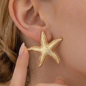 Könnte beinhalten: Goldene Seestern-Ohrstecker. Die Ohrringe sind strukturiert und haben ein detailliertes Design. Die Ohrringe werden am Ohr getragen. Die Ohrringe sind ein Accessoire zum Thema Strand.