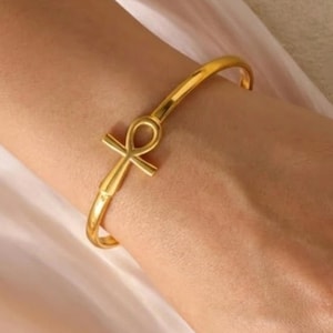 Puede incluir: Una pulsera dorada con un diseño de cruz Ankh. La pulsera es de estilo brazalete abierto y sencillo, y el Ankh está centrado en la parte delantera. La pulsera se lleva en una muñeca y el fondo es de color neutro.