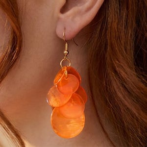 Puede incluir: Pendientes de disco naranja con ganchos y anillos de conexión dorados. Los pendientes presentan múltiples discos circulares naranjas superpuestos que caen en cascada. Los pendientes se muestran sobre un fondo de cabello y piel de color marrón rojizo.