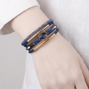May include: A multi-strand bracelet set featuring blue beads, gold-toned metal accents, and a central oval blue stone. The bracelets are stacked on a wrist, showcasing the design and color contrast.