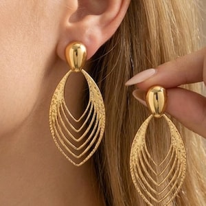 Könnte beinhalten: Goldfarbene Ohrringe mit einem strukturierten, geschichteten Design. Die Ohrringe haben eine Tropfenform mit mehreren absteigenden, gebogenen Linien. Der obere Teil des Ohrrings ist ein glatter, abgerundeter Stecker.