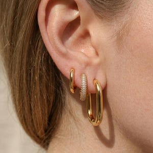 Könnte beinhalten: Nahaufnahme eines Ohrs mit drei goldenen Ohrringen. Die Ohrringe umfassen einen kleinen, einfachen Reifen, einen mit kleinen, funkelnden Steinen besetzten Reifen und einen größeren, rechteckigen Reifen. Die Ohrringe haben eine warme, glänzende Goldfarbe.