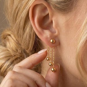 Könnte beinhalten: Goldene Ohrringe mit einem kleinen Kugelstecker und drei hängenden Ketten, an denen jeweils eine goldene Kugel befestigt ist. Die Ohrringe werden von einer Person mit blondem Haar getragen. Schlichtes, elegantes Design.