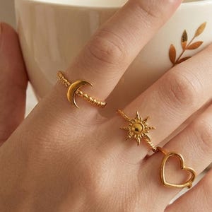 Könnte beinhalten: Goldfarbene Ringe mit himmlischen und romantischen Designs. Ein Ring hat einen Halbmond, ein anderer einen Sonnenaufgang und der dritte eine Herzform. Die Ringe werden an Fingern getragen, mit einer weißen Tasse im Hintergrund.
