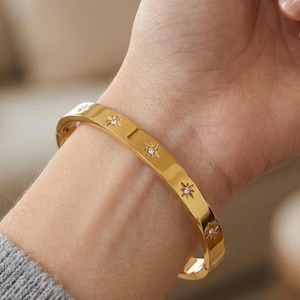 May include: A gold-coloured bangle bracelet featuring star-shaped details, each set with a small, clear gemstone. The bracelet is worn on a wrist, and the design is simple and elegant. The bracelet is a piece of jewellery.