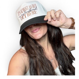 May include: A white trucker hat with a black brim featuring the text IT'S ALMOST AS HOT as my husband OUT HERE printed in a mix of black and copper-coloured lettering on the front panel of the cap.