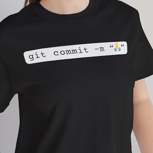May include: Black t-shirt featuring the text "git commit -m "" in white. The top has a crew neck and short sleeves. The text includes a code-like command with a small emoji.