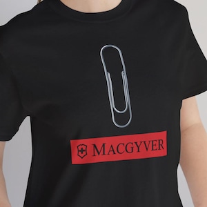 Könnte beinhalten: Schwarzes T-Shirt mit einer silbernen Büroklammer und dem Wort "MACGYVER" in Weiß auf einem roten Rechteck. Das T-Shirt hat einen Rundhalsausschnitt.