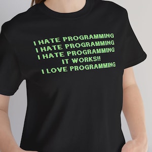 Może przedstawiać: Czarny t-shirt z tekstem "I HATE PROGRAMMING" powtórzonym trzy razy, a następnie "IT WORKS!!" i "I LOVE PROGRAMMING" w kolorze zielonym. Koszulka ma okrągły dekolt.