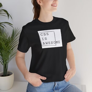 Op de afbeelding: Zwart T-shirt met de tekst "CSS IS AWESOME" in een witte rechthoek. Het T-shirt heeft een ronde hals en wordt door een persoon gedragen. De persoon draagt een blauwe jeans.