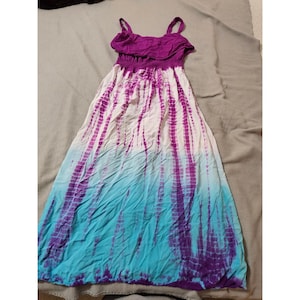 May include: A tie-dye maxi dress with a purple ruffled top and spaghetti straps. The dress transitions from white to purple and then to turquoise. The fabric is gathered at the waist and flows down to the floor.