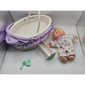 May include: A toy bassinet with a purple fabric exterior and a white quilted interior, featuring a white handle. Beside it lies a doll wearing a white sleeper with a purple and blue floral pattern, along with a small green plastic brush and bottle.