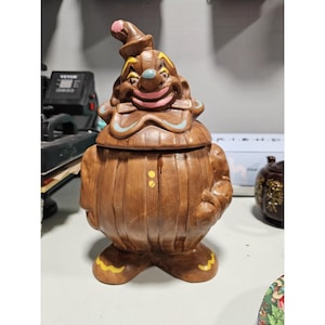 May include: A brown ceramic clown cookie jar with a smiling face, a pink nose and mouth, and a pointed hat. The jar has a barrel-shaped body with yellow accents on the feet.