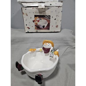 Puede incluir: Un cuenco de cerámica blanco para caramelos con forma de muñeco de nieve, con sombrero amarillo, bufanda burdeos y botas negras. El muñeco de nieve sostiene un cartel de "Se busca" y un aperitivo. El cuenco viene con una caja decorativa.