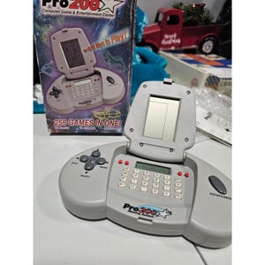 以下が含まれることがあります： グレーのPro 200ゲームシステム。フリップアップスクリーンとキーパッド付き。「Computer Game & Entertainment Center」と「256 Games in One!」と書かれた箱が後ろにあります。システムには方向パッドと回転/モードボタンがあります。