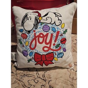 Puede incluir: Cojín cuadrado blanco con Snoopy con gorro de Papá Noel, descansando sobre una corona con adornos coloridos y la palabra "joy!" en rojo. Un gran lazo rojo está en la parte inferior. Decoración festiva navideña.