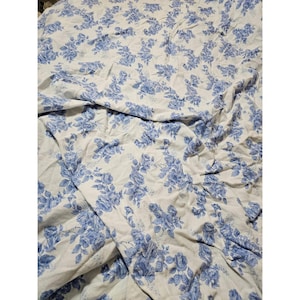 May include: A white fabric sheet featuring a repeating blue floral pattern of roses and leaves. The material is draped in soft folds, displaying the intricate botanical print across the entire surface of the textile in a consistent design.