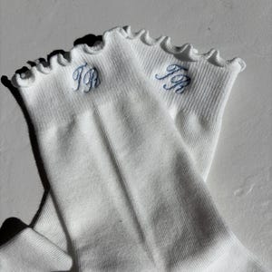 Op de afbeelding: Een paar witte geribbelde sokken met een gefranjerde bovenrand. De sokken hebben lichtblauwe geborduurde initialen. De sokken zijn gemaakt van een zacht, comfortabel materiaal.