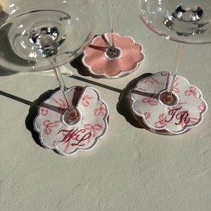 Puede incluir: Tres colgantes para copas de vino con diseño floral e iniciales bordadas. Los colgantes son blancos con estampados florales rosas y bordes festoneados. Un colgante es rosa liso. Las iniciales "WL" y "TR" están bordadas en rojo.