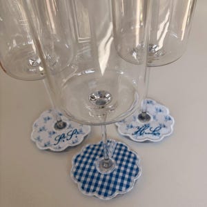 May include: Clear wine glasses with decorative coasters. The coasters feature floral and gingham patterns in blue and white, with initials embroidered on some. The glasses are arranged on a white surface.