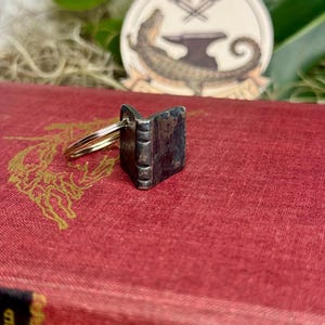 May include: A small, silver-toned book-shaped keychain. The miniature book is open, revealing the pages. The keychain is resting on a red book with gold lettering. A round sticker with an alligator and anvil design is in the background.