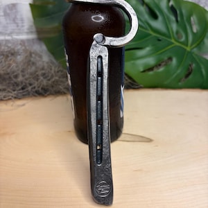 May include: A dark gray, hand-forged metal bottle opener shaped like a horseshoe. The tool features a curved hook at the top for gripping bottle caps and a straight, textured handle with a small circular stamp at the bottom end, showing a hammer icon.