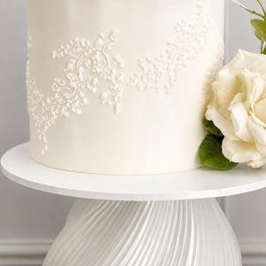 Op de afbeelding: Een witte meerlagige taart met delicate bloemendetails en een grote witte roos. De taart staat op een witte sokkel met een textuur, wervelend ontwerp, wat zorgt voor een elegante presentatie.