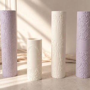 May include: Four cylindrical vases in a neutral setting. Two are a soft lavender, and two are off-white. All feature a raised floral pattern. The vases vary in height, with the shortest being about 15 cm tall.