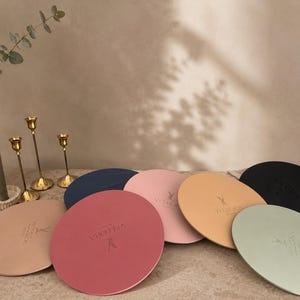 Könnte beinhalten: Eine Auswahl an runden Tischsets in verschiedenen Farben, darunter Rosa, Pfirsich und Schwarz, ist auf einem Tisch angeordnet. Jedes Tischset hat ein Logo. Dekorative Kerzen und goldene Kerzenhalter sind im Hintergrund.