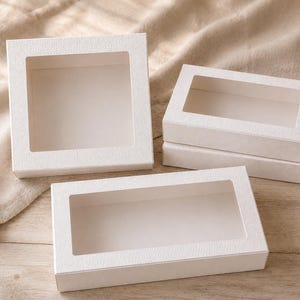 Peut inclure: Plusieurs boîtes en carton blanc texturé avec des découpes fenêtres transparentes. Une boîte est carrée, les autres sont rectangulaires. Les boîtes sont disposées sur une surface claire et texturée, pour l'emballage ou la présentation.