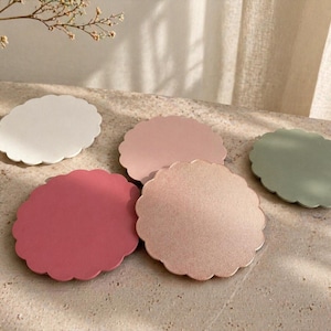 Può includere: Cinque sottobicchieri rotondi con bordo smerlato nei toni del bianco, rosa e verde. Sono disposti su una superficie strutturata. Un vaso con fiori secchi è sullo sfondo. I sottobicchieri sono ideali.