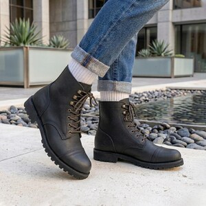 Puede incluir: Un par de botas de cuero negro con cordones y puntera reforzada, con ojales de metal oscuro, cordones marrones y una suela de goma gruesa con tacos, combinadas con pantalones vaqueros azules y calcetines blancos de canalé hasta el tobillo.