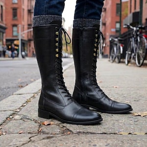 May include: Black knee-high boots with a lace-up design and gold eyelets. The boots have a side zip and a low heel. The boots are worn with blue jeans and grey socks.