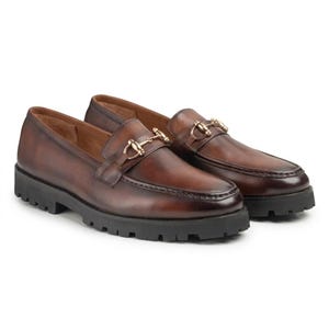 Handmade brown leather horsebit loafer with cuffed jeans on cobblestone path with autumn leaves
