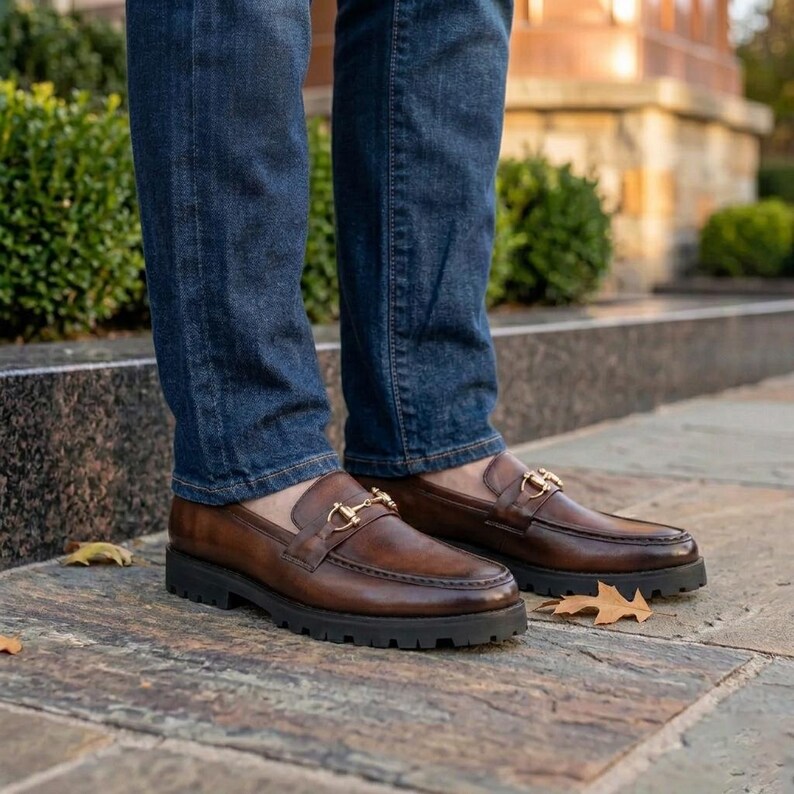 Angled view of brown men's leather loafer showing gold buckle strap and black lug sole on white background