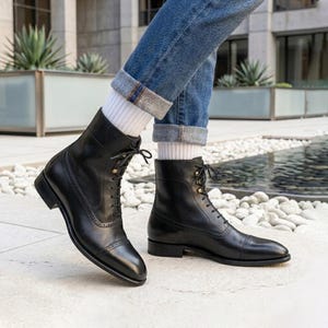 May include: A pair of black leather lace-up boots featuring a cap toe with decorative brogue detailing, metal eyelets, and thin black laces. The boots are worn with white ribbed socks and blue denim jeans with rolled cuffs at the ankles.