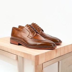 Op de afbeelding: Een paar bruine leren herenschoenen met een klassiek ontwerp. De schoenen hebben een gepolijste afwerking, een gestikte zool en bruine veters. De schoenen worden gepresenteerd op een lichtgekleurd houten oppervlak.