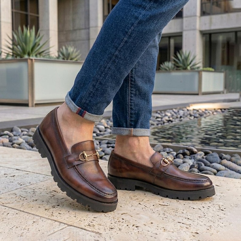 Mens dark brown burnished leather horsebit loafer with gold buckle worn with jeans on autumn stone steps