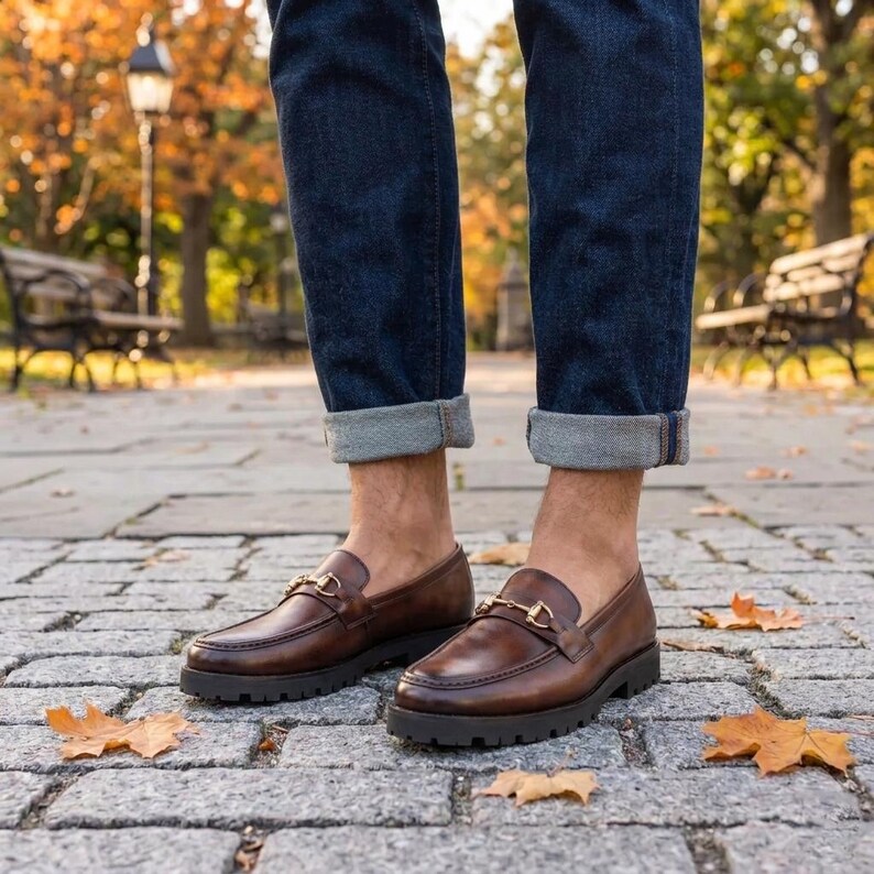 Mens brown leather lug sole loafer with gold horsebit worn with rolled denim near outdoor water feature