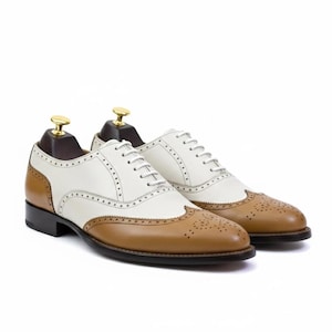 Può includere: Un paio di scarpe stringate bicolore. Le scarpe presentano una tomaia bianca con dettagli marroni sulla punta, sul tallone e sui pannelli laterali. Design classico con lacci.