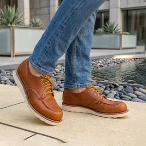 May include: A pair of brown leather oxford shoes with moccasin-style stitching on the toe, white rubber soles, and yellow laces. The shoes are worn with blue denim jeans, showing a low-top silhouette with metal eyelets and visible white stitching.