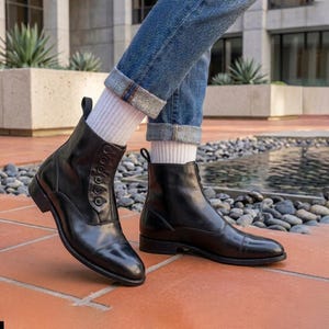 May include: A pair of black leather ankle boots with button detailing and a zipper closure. The boots are worn with blue jeans and white socks. The boots have a classic design and a polished finish, suitable for various occasions.